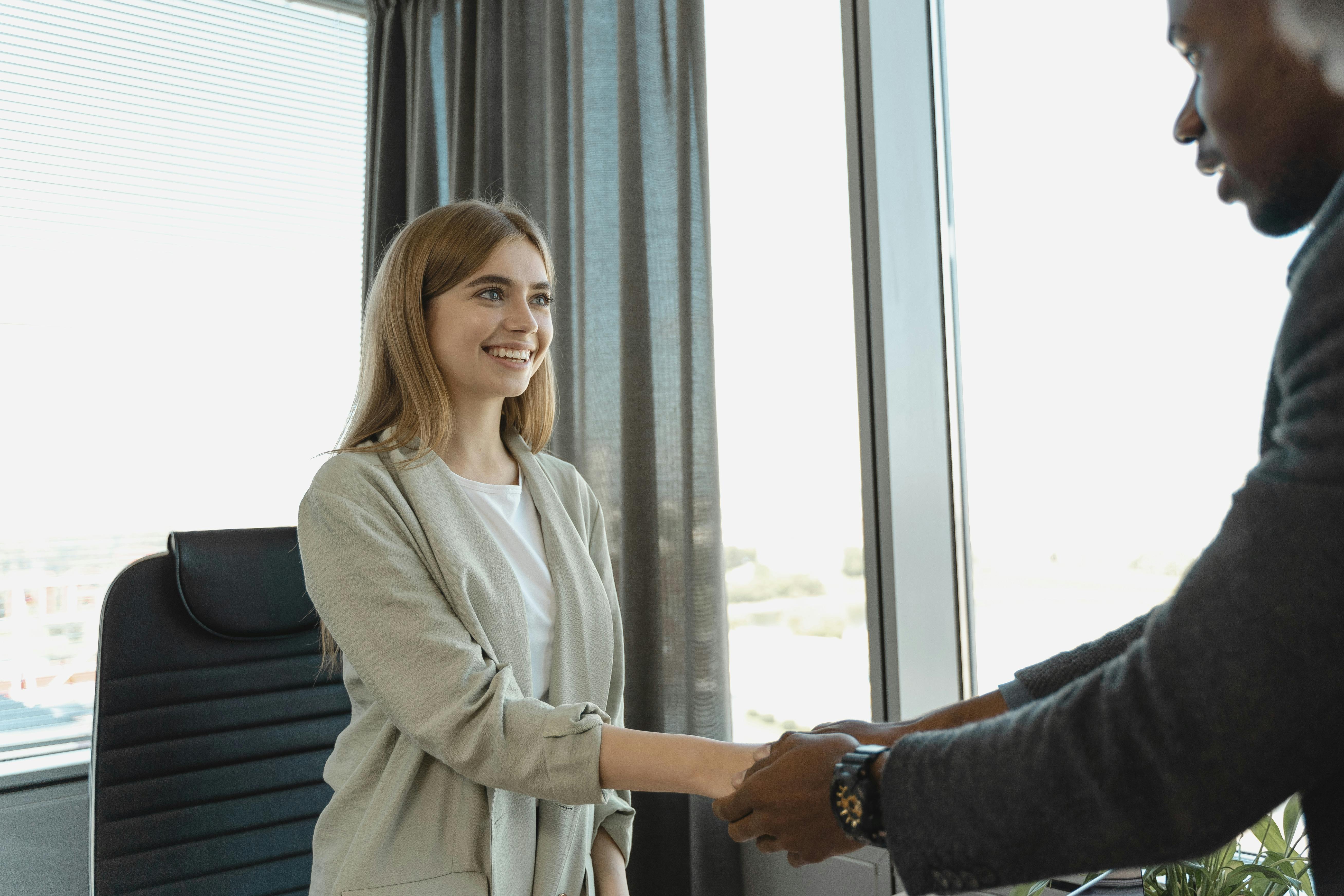 Young female shaking hands with a potential employer at a job interview. Female is wearing a beige jacket and white tshirt.